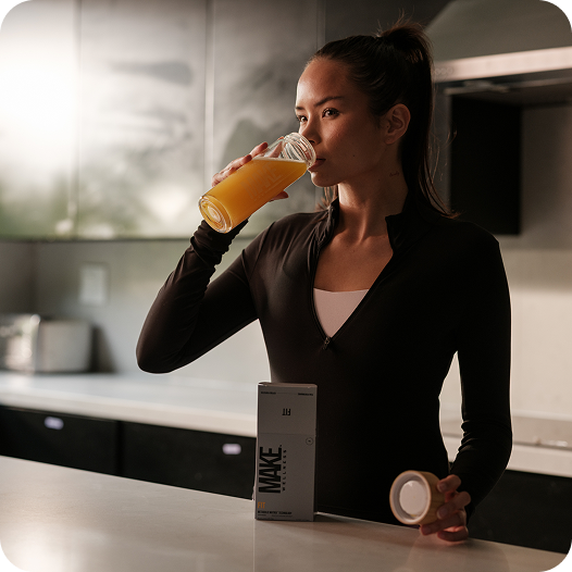Woman drinking MAKE peptide drink in dark kitchen, MAKE box visible on counter